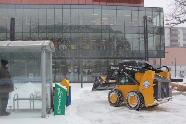 Plow clears snow in front of Halifax Central Library while someone observes.