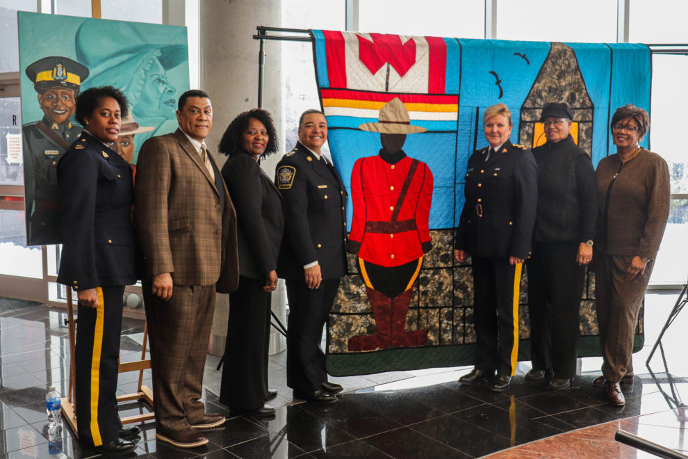Attendees at the Feb. 13 RCMP event for "Her Story" pose with a painting and quilt made to commemorate black canadians in law enforcement.