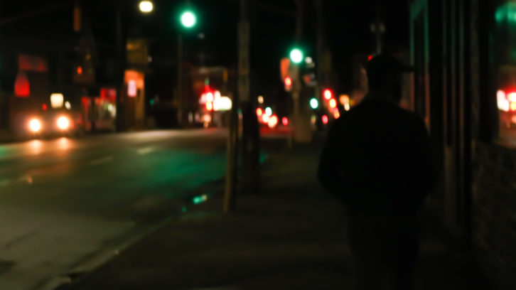 A landscape street view of Quinpool at night, with one car. Silhouette of a man on the right side of the image.