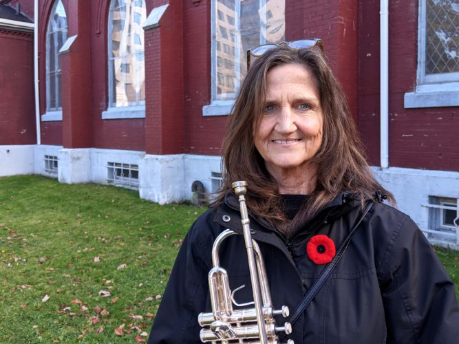 Sue Mantin stands with her trumpet outside a St. Mark's Anglican church. She wears a black jacket and a red poppy.