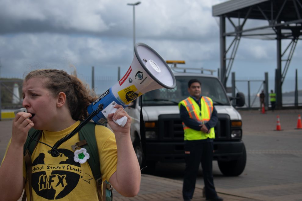 Organizer Willa Fisher leads a chant against Nova Scotia Power as a security personnel looks on, outside their downtown Halifax headquarters on Lower Water Street
