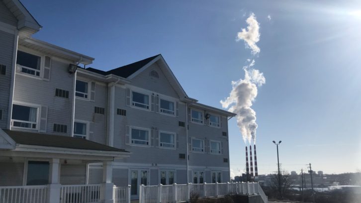 A large white hotel fills the frame. In the background are blue sky and three tall red and white chimneys pumping out a cloud of white smoke.