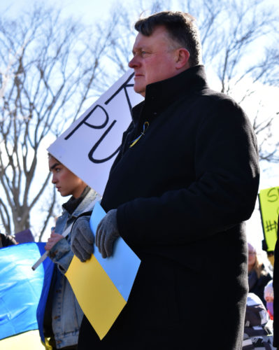 Halifax Mayor Mike Savage stares to the left while wearing a black jacket and holding a yellow and blue placard.