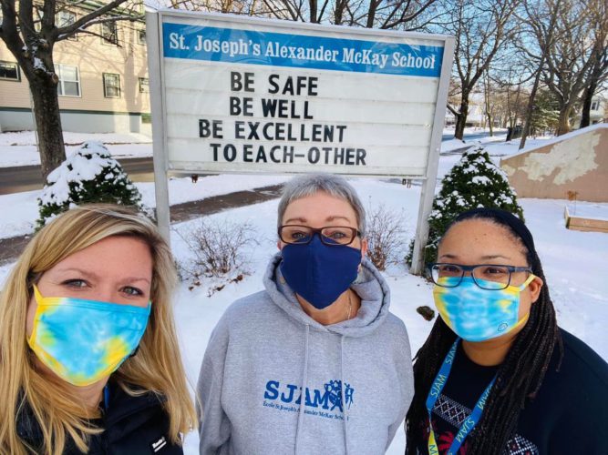 Three women pose in front of a sign that says "Be safe, be well, be excellent to each other."