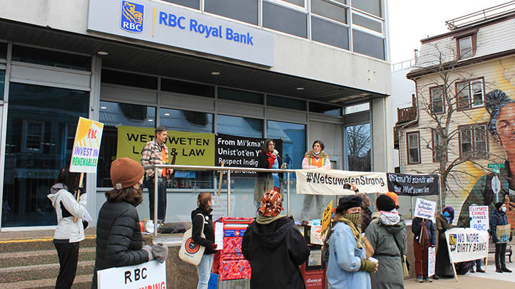 16 people gathered in front of an RBC. 3 stand on a ledge being used as a stage. They are surrounded by banners with slogans like “#WetsuwetenStrong,” “Mi’kma’ki stands with the Wet'suwet'en”