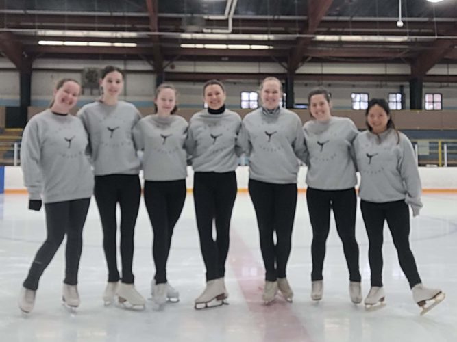 Seven women stand in a line in an ice rink, wearing grey shirts, black leggings and figure skates