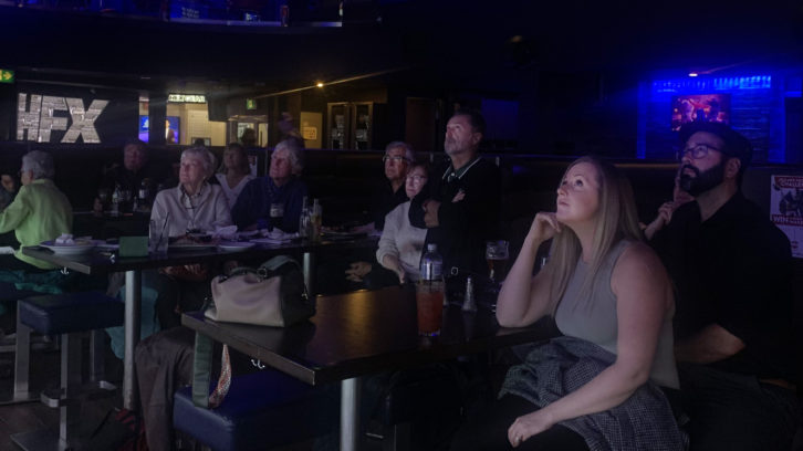 Nine people. Sat around tables in a dimly lit bar, gazing upwards and watching the “Jeopardy” match (not pictured). A light up sign which reads “HFX” glows in the background.