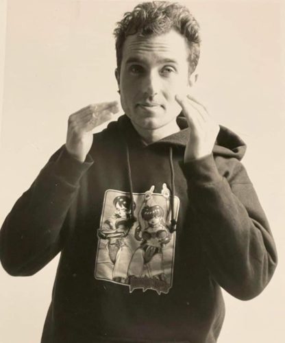 A black and white photo of Chris Redden. He has wavy, short hair, a stud earring, and is wearing a hoodie with anime characters on it. He has his hands raised in mid-motion.