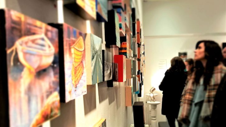 A person in the crowd stands and looks at paintings on the wall.