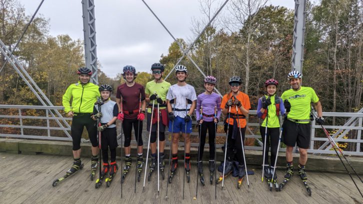 The N.S. XC team is lined up on a bridge. They are all wearing helmets and roller skis.