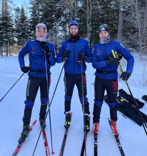 Teammates Lyndon Doyon, Cohen Norman and Jack MacMillan stand together with their skis on. They are wearing blue team jackets and black pants. They are all holding poles and wearing buffs with glasses resting on their heads. Behind them are trees and a few black ski bags laying in the snow.