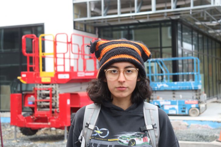 Young woman with glasses and knit hat poses for picture outside a construction project.