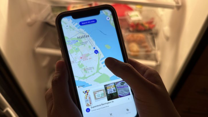 An iPhone is open to the FlashFood app, which shows a map of Halifax and the discounted items available at the nearest superstore. The phone is held by two hands, and lit up from behind by an open refrigerator.