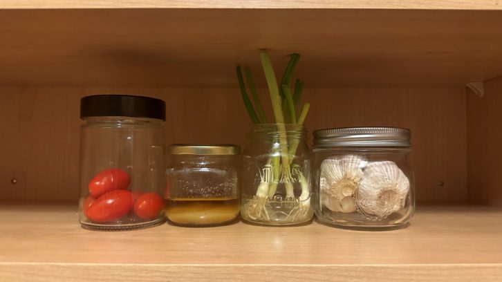 A cupboard shelf with four ingredients lined up on it. From left to right: a jar containing about a third of cherry tomatoes, a smaller jar containing used cooking oil, a jar filled with water from which young green onions are growing, and a round jar containing bulbs of garlic.