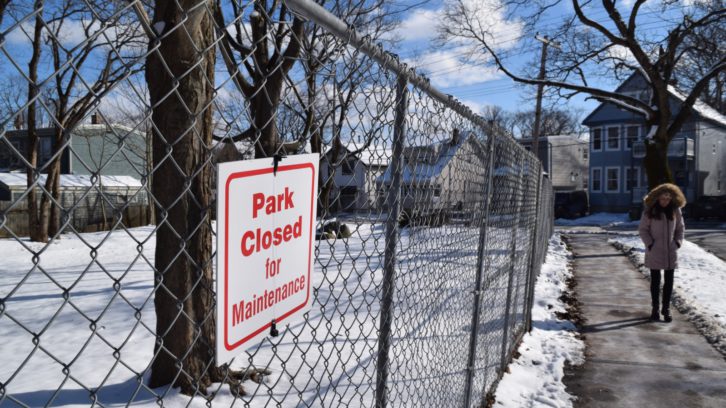 Woman in pink winter jacket walks by the fenced-off, snow-covered People’s Park and a white sign that reads in red letters: “Park closed for maintenance.”
