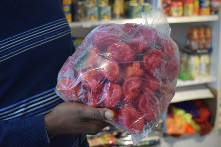 Hands hold out a bag of frozen red chilli peppers called “Scotch bonnets.” The man holding them is wearing a navy blue, striped sweater. There are shelves of cans and dry goods in the background.