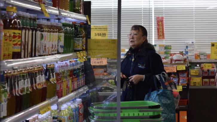 An older woman with black hair and glasses walks by an open fridge stocked with drinks as she carries grocery bags on her arm and keys in her hands. There are yellow and orange signs surrounding her with prices of items in Union Foodmart’s Dartmouth location. She wears a dark blue, zip-up sweater.