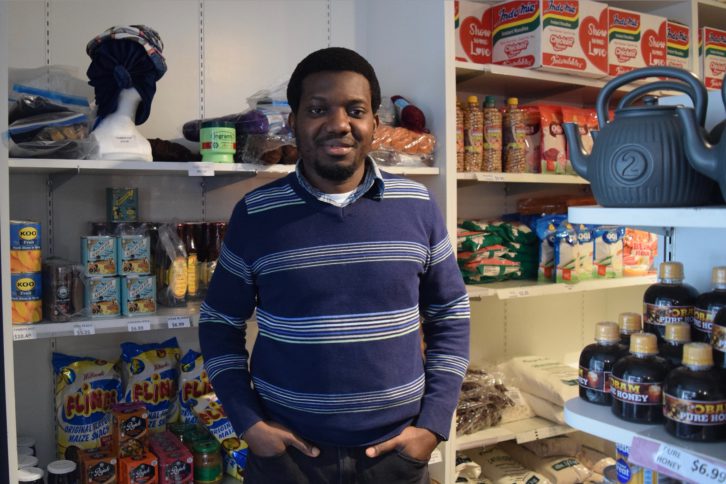 Man poses for the camera with hands in the pockets of his black jeans. He wears a navy-blue sweater with small stripes. Behind him are African products like chips, canned fruit and rice.