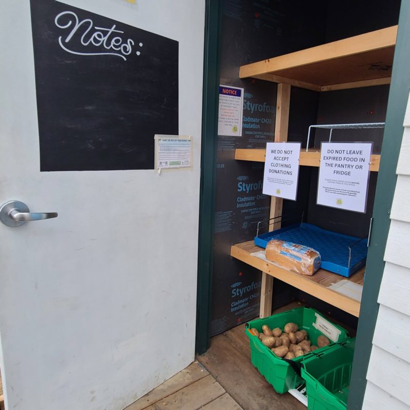 The pantry door of the Dartmouth Community Fridge is open, showing the shelves and notices inside. There is a loaf of bread and a box of potatoes visible. There is a note board on the door.