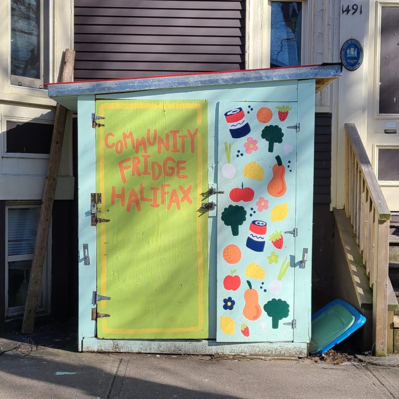 The left-hand side fridge door of the Halifax Community Fridge shows the painted words "community fridge Halifax". It is sunny outside.