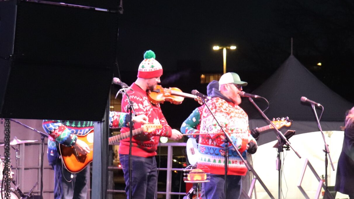 Four band members wearing holiday sweaters and hats are performing on stage. Two members are playing the guitar, one is playing the fiddle, and another is singing.
