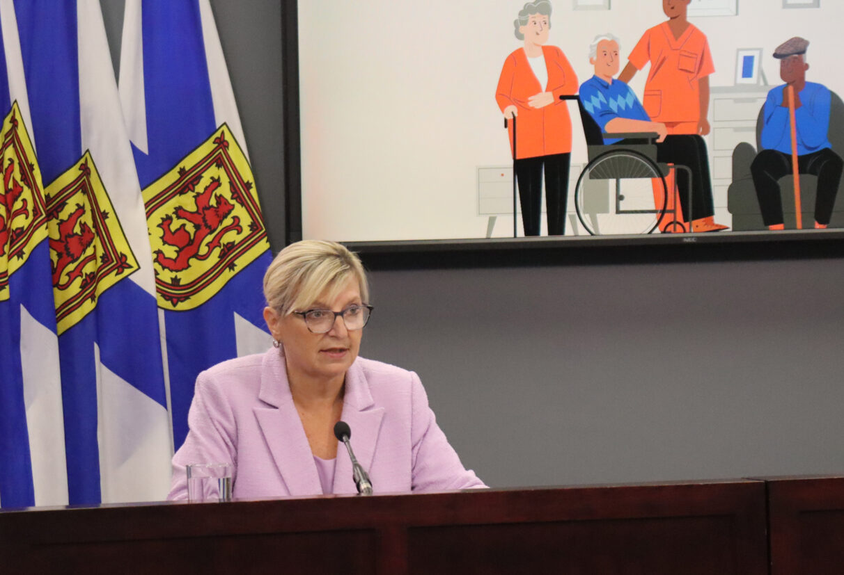 A woman sitting at a desk with Nova Scotia flags and a projector with healthcare pictures behind her.
