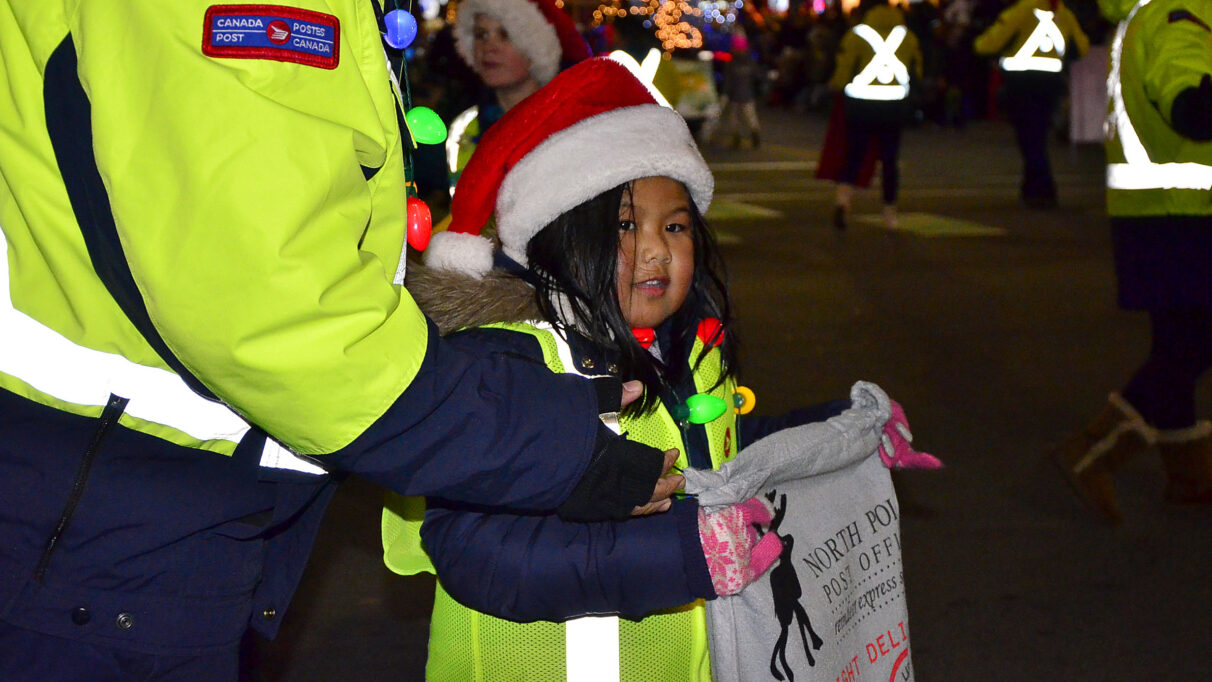 A child wearing a neon yellow vest and a Santa hat holds a bag collecting letters from kids for Santa.