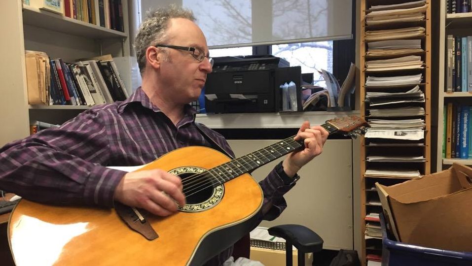 Professor Jason Brown playing guitar in his office.