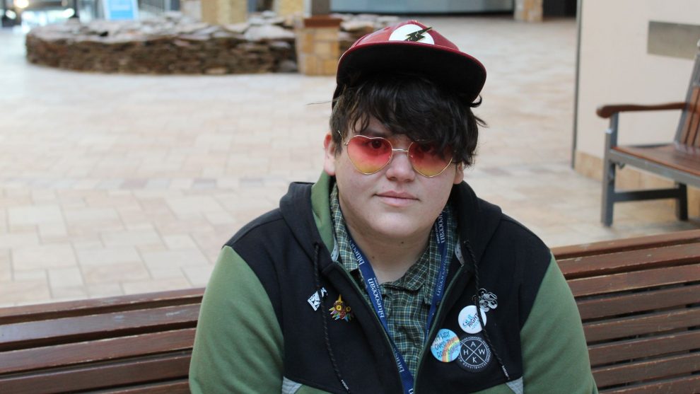 Jace Miller sits on a bench wearing heart-shaped sunglasses and buttons on his sweater. One reads, "I'm way over the rainbow."