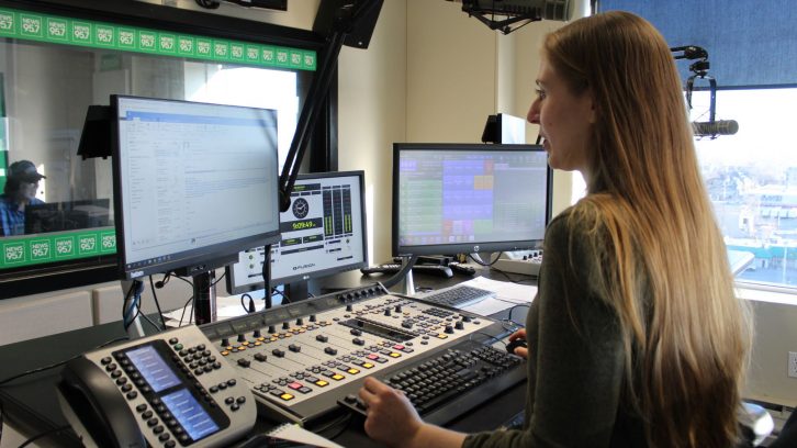 Katie Hartai stands at her desk in the radio room at News 95.7. In the background Rick Howe can be seen talking on the radio.