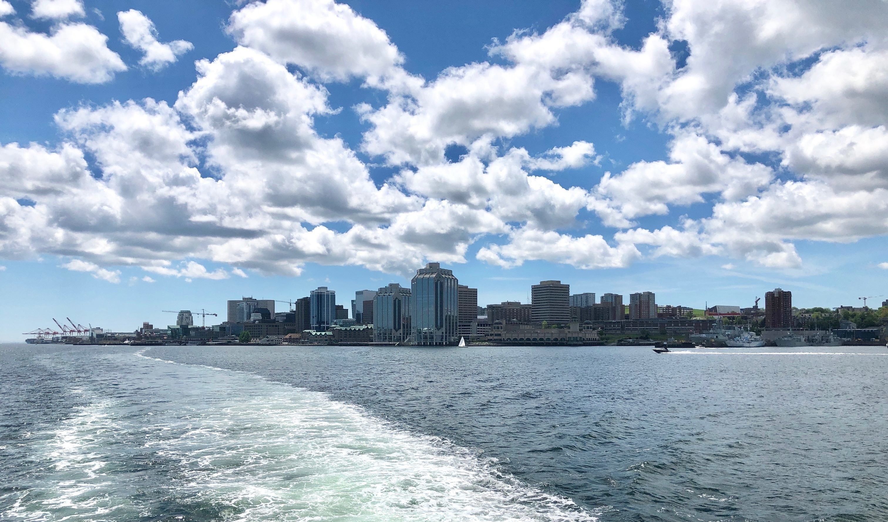 An image of the city skyline of Halifax, Nova Scotia. The buildings are framed by blue water and a cloudy blue sky.