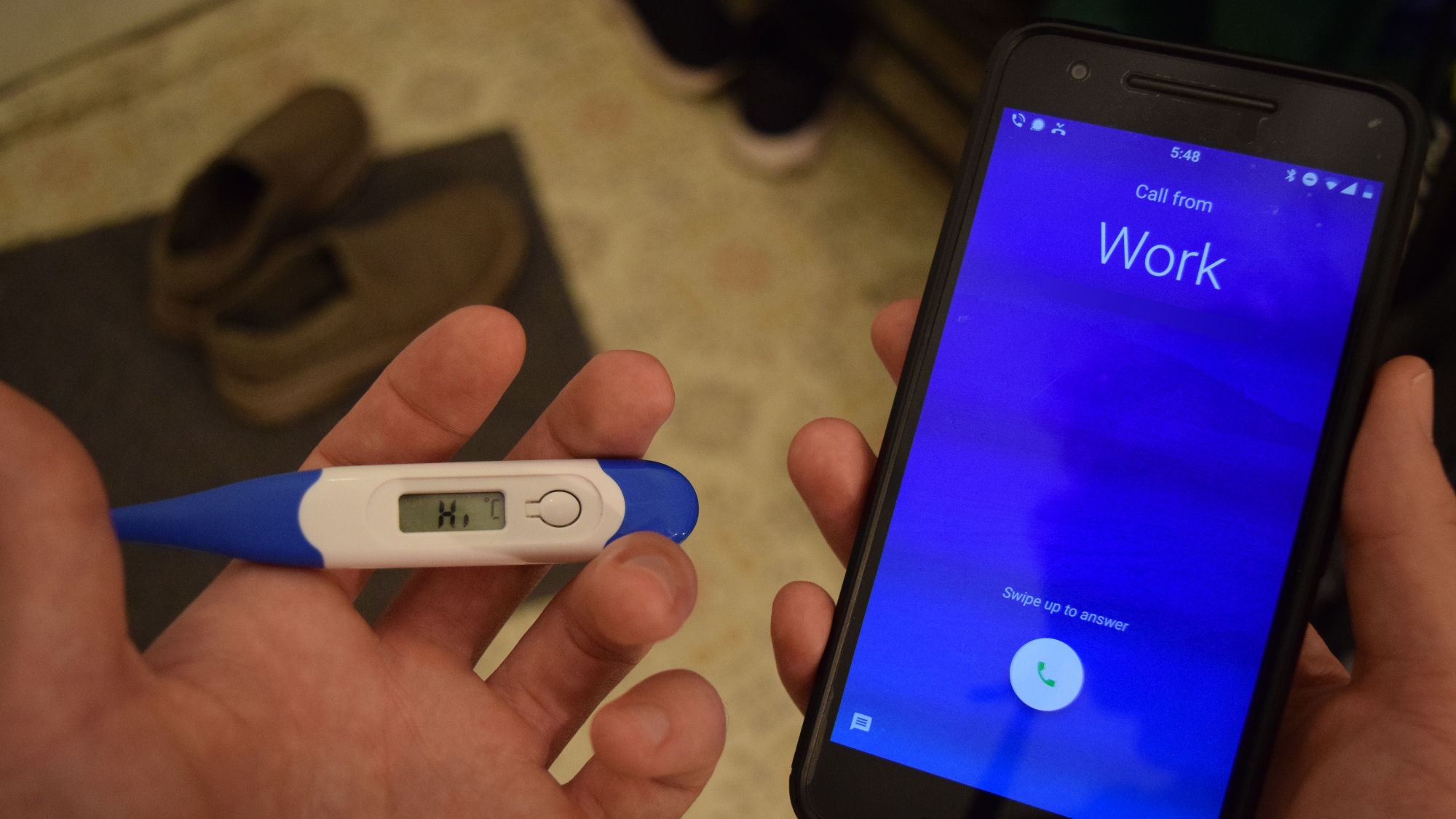 Someone holds a thermometer with a high temperature reading in one hand and a phone receiving a call from work in their other.