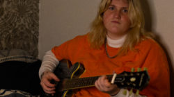 Em Burry sits on their bed while holding their guitar. They are a young artist carving their path in a shaken industry.