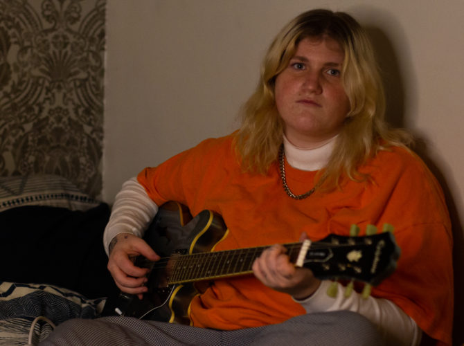 Em Burry sits on their bed while holding their guitar. They are a young artist carving their path in a shaken industry.