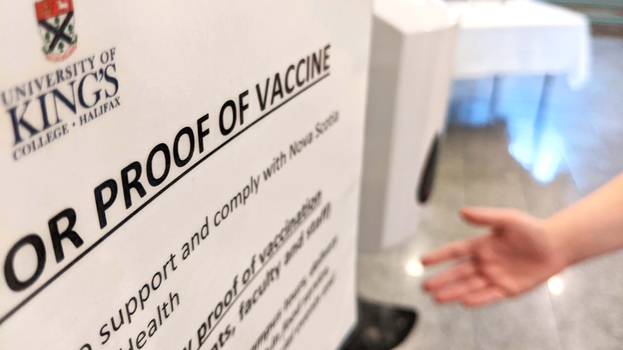 A proof of vaccination requirement sign sits in the foreground while a hand reaches for sanitizer behind it.