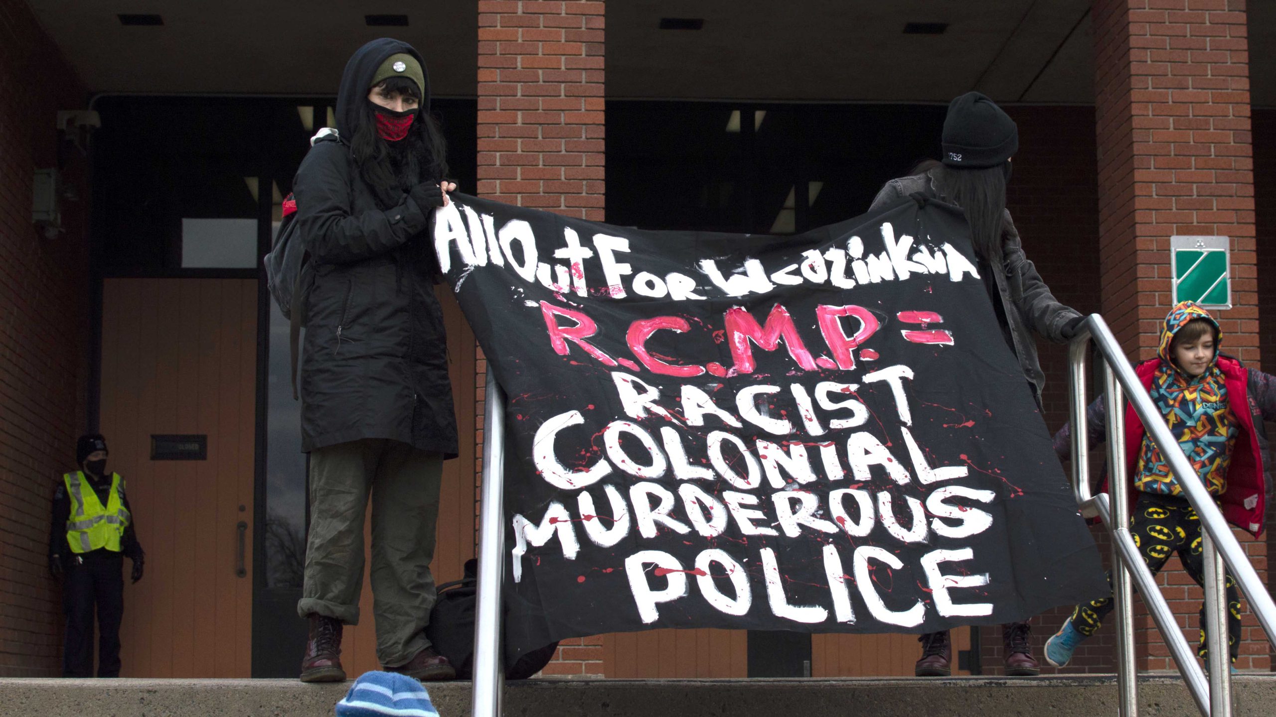 A police officer guards the entrance while demonstrators hold a sign that says "All out for Wedzin Kwa / RCMP = racist, colonial, murderous police" on the steps of the Halifax Regional Police headquarters on Nov. 21.