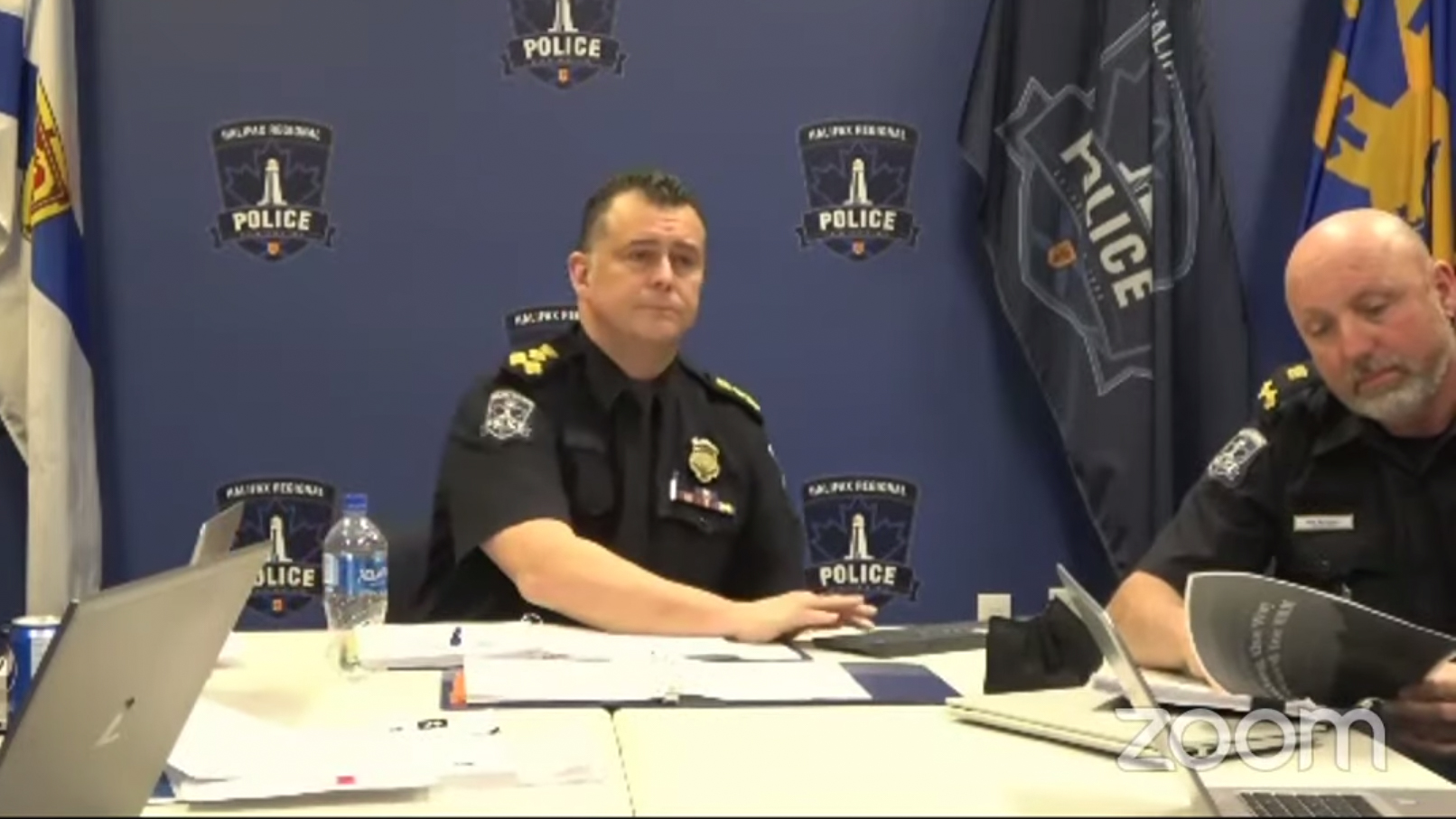 A Zoom screenshot of Halifax Regional Police Chief Dan Kinsella and Deputy Chief Reid McCoombs sitting at a table in front of a blue wall with HRP logos. McCoombs is looking through what appears to be the subcommittee's report on defunding the police.