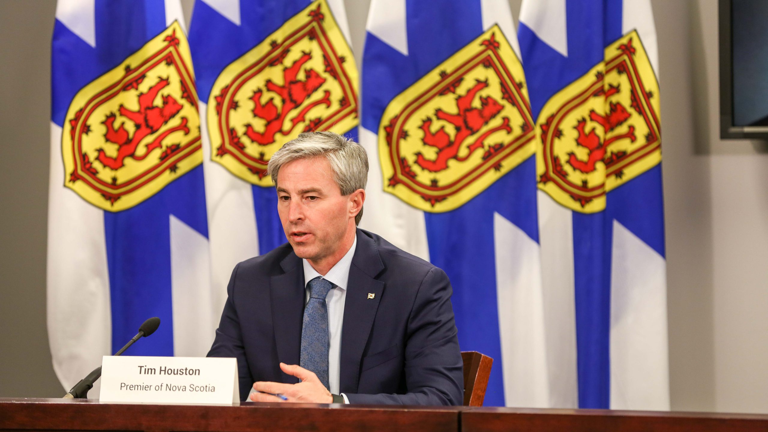 Premier Tim Houston sits at a desk in front of four Nova Scotian flags while speakings.