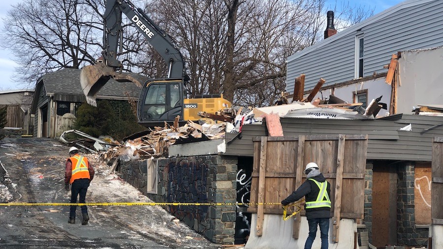 A construction worker drags yellow tape across the entrance to a demolished property. There is wood, stone and debris everywhere and a tractor crane in the background.