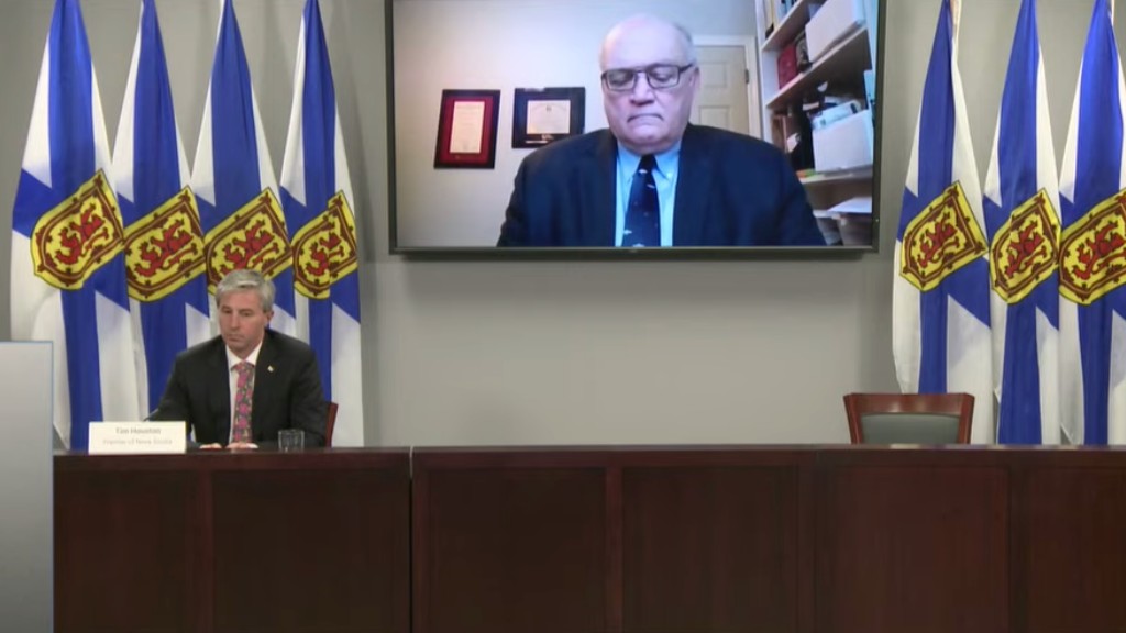 Premier Tim Houston sits at a large desk. Dr. Robert Strang is projected onto a screen above him.