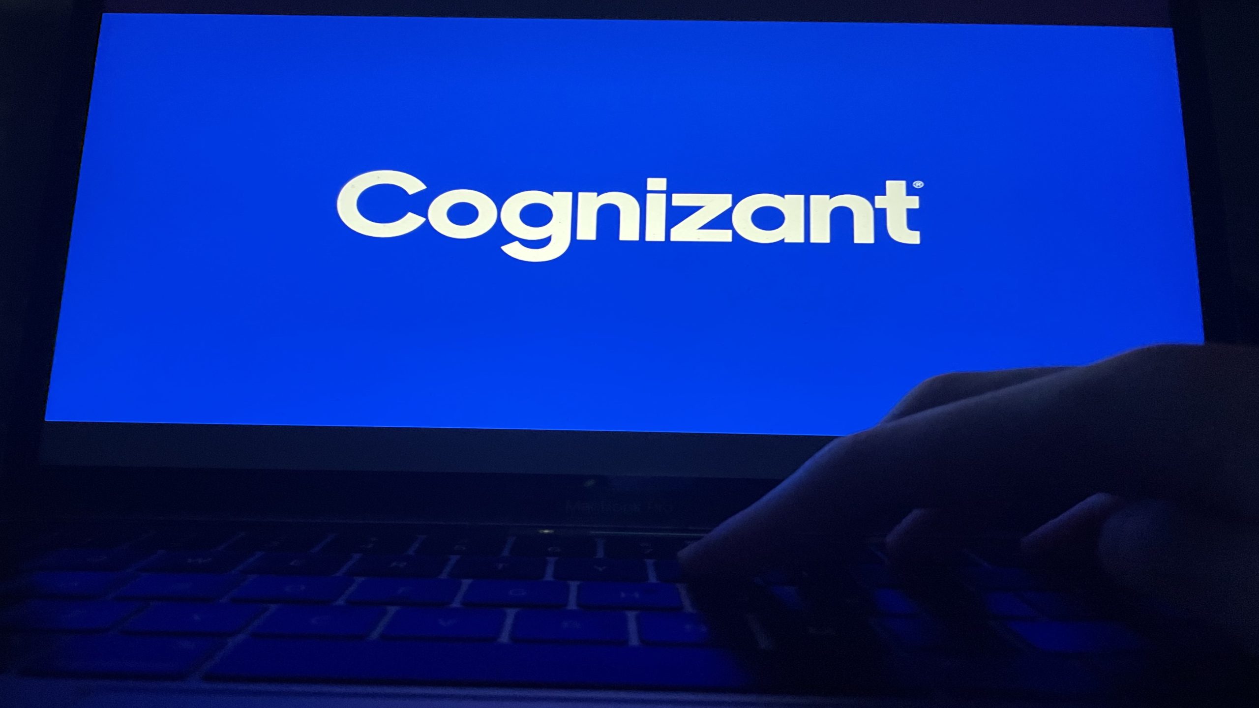Bathed in blue light, a hand works on a laptop glowing with the Cognizant logo