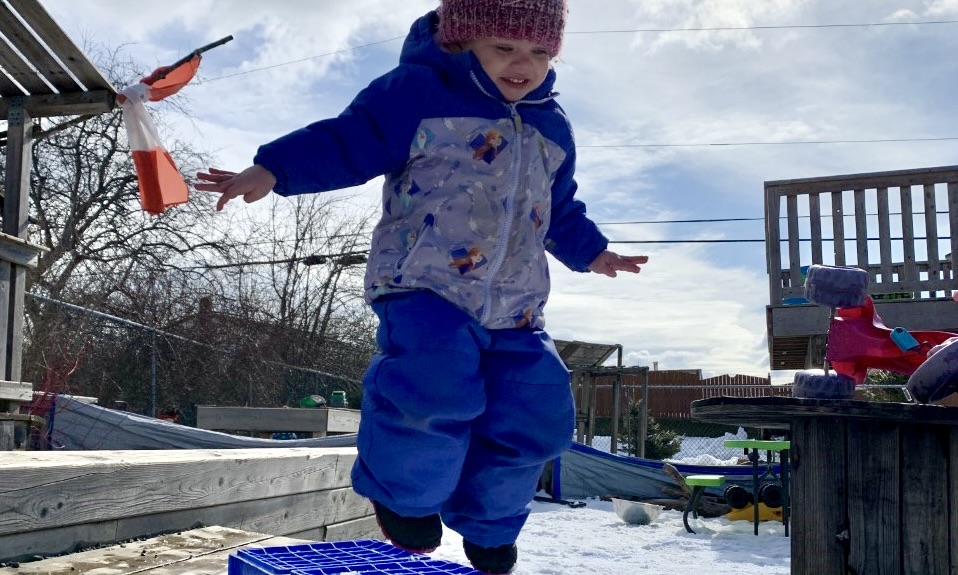 A toddler dressed in winter clothing jumps off of a blue crate into the snow.