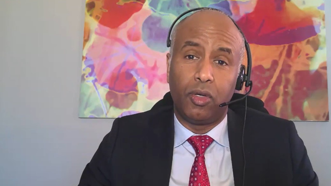 A man wearing a headset speaks in a screenshot from a virtual press conference. There is a colourful art piece in the background.