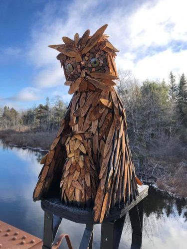 A large wooden owl sculpture with pennies for a face sits atop a metal frame with a blue sky in the background and water behind it.
