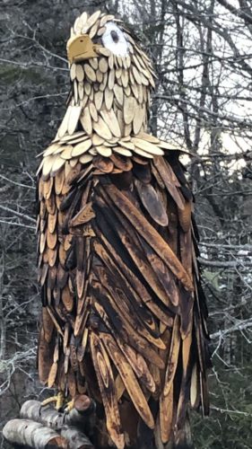 A wooden eagle sculpture perches on logs on top of a metal frame with trees in the background.