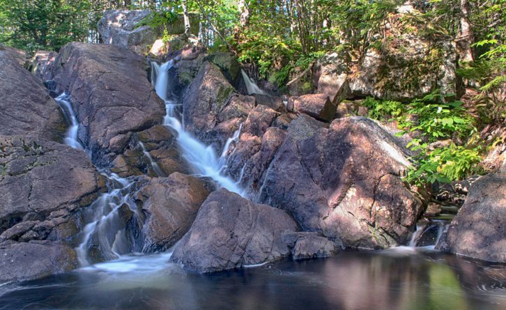 A low-volume waterfall with rocks exposed sits above a dark pool surrounded by woods. 