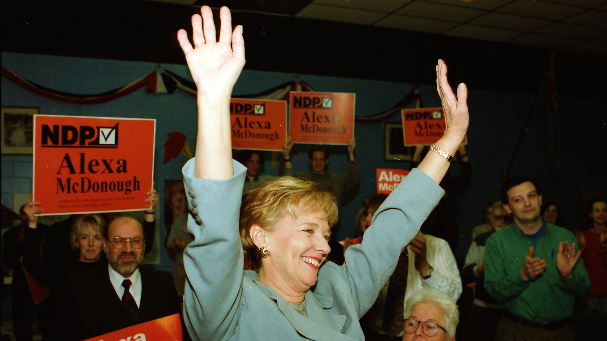 Alexa McDonough raises her arms in victory while surrounded by supporters with signs.