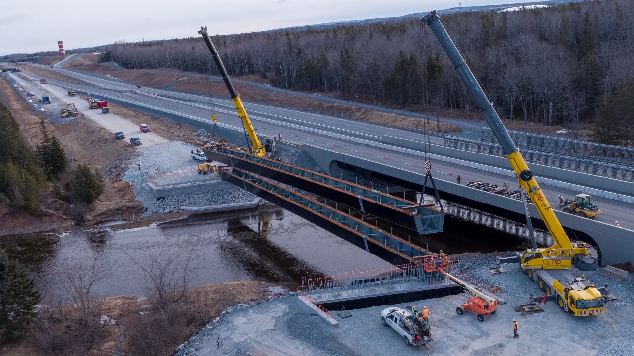 Two large cranes are seen on either side of a river. They are lifting a section of bridge into place.
