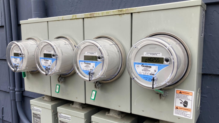 Four electric meters are are seen side by side, mounted on metal boxes with an eelctric shock warning sticker in the lower right.