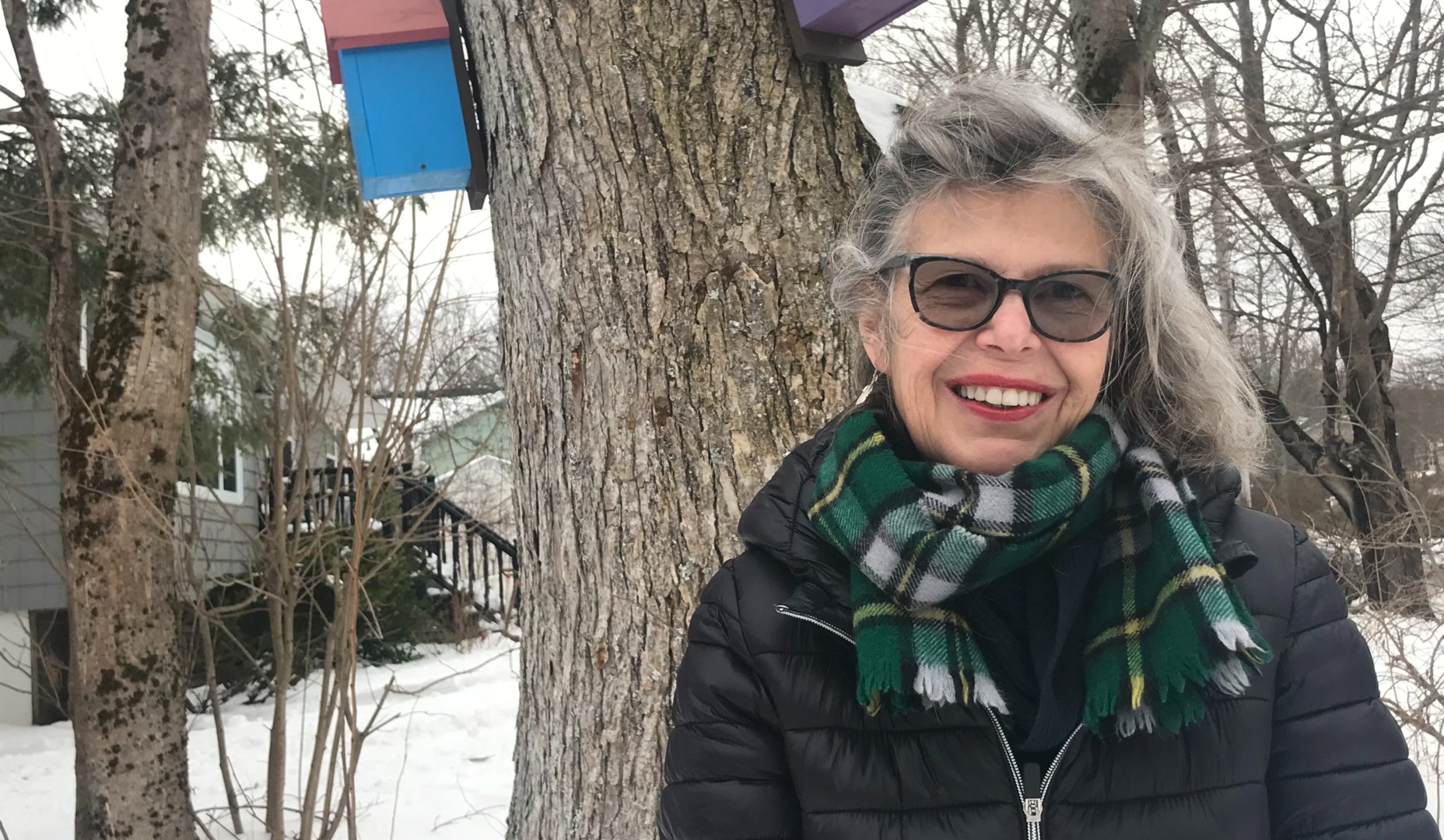 A woman with grey hair and a green scarf poses in front of a tree on a snowy day.